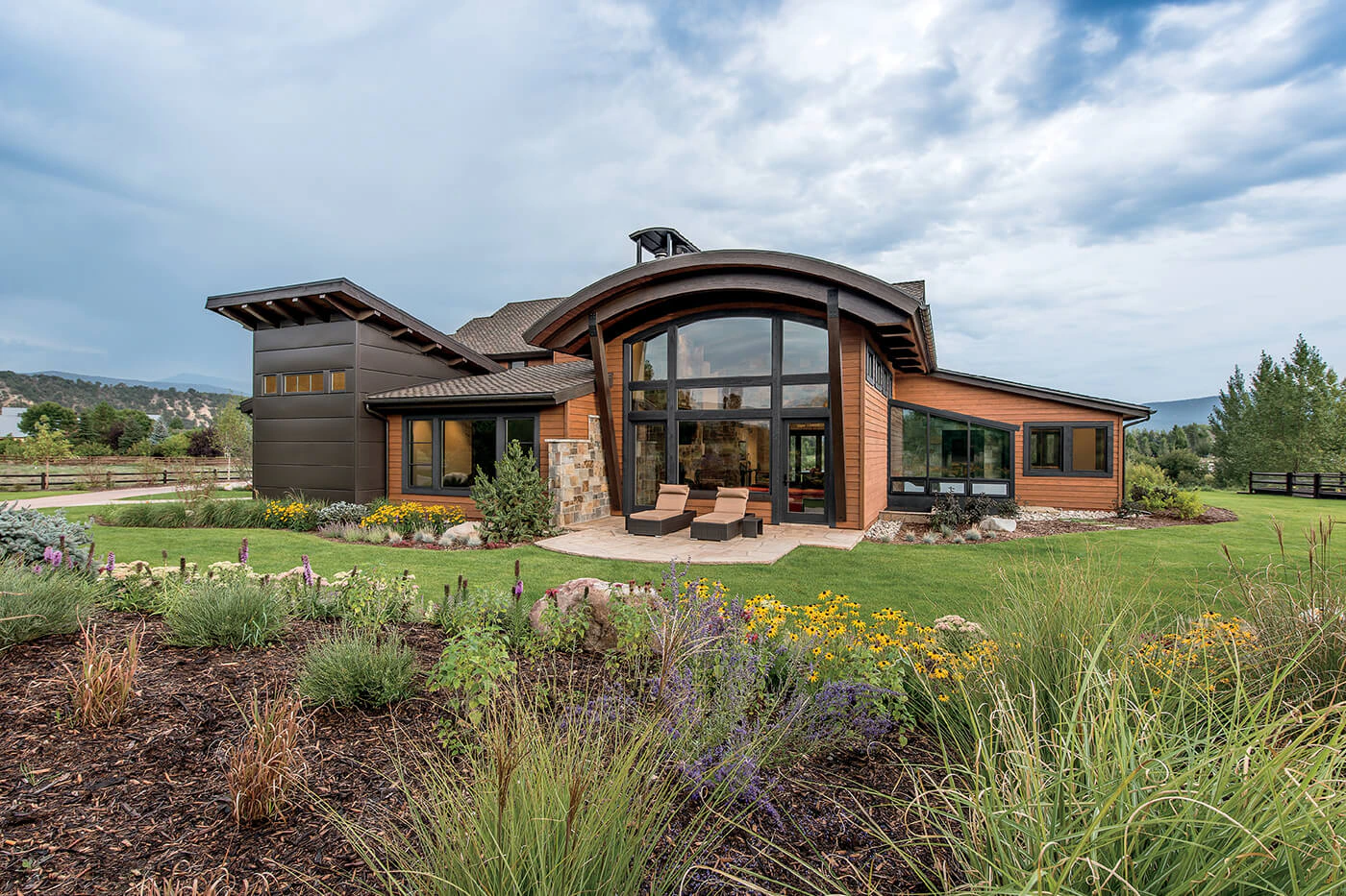 Modern mountain-style home with large arched windows, wood and metal exterior, surrounded by landscaped garden with colorful flowers and lush green lawn under a cloudy sky.